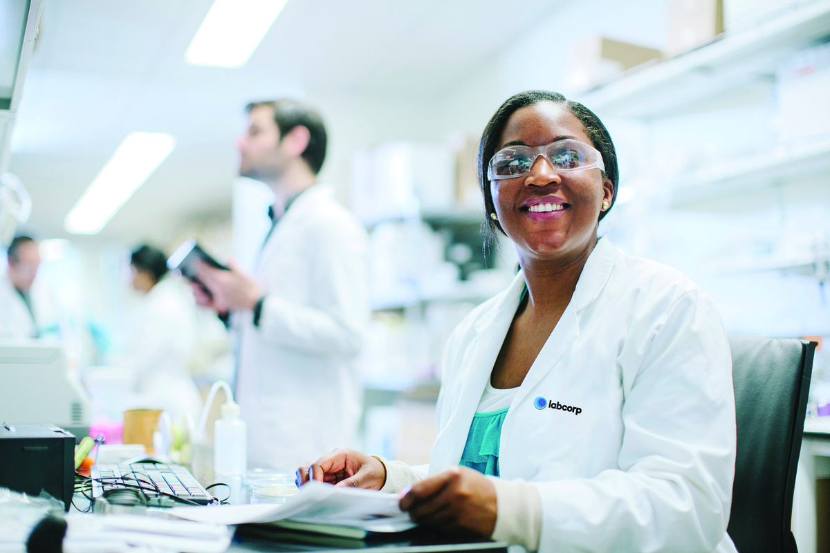 Scientist sitting at a desk in a lab. She is wearing goggles and has a big smile