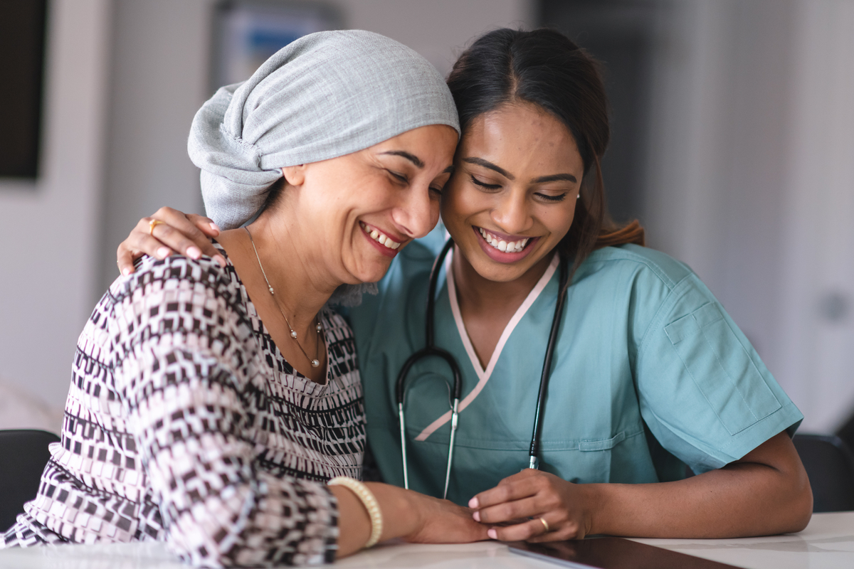 1175131167 Portrait of an Indian woman fighting cancer with her physician. The doctor is a mixed-race female. The two women are seated next to each other indoors. They are embracing and laughing.