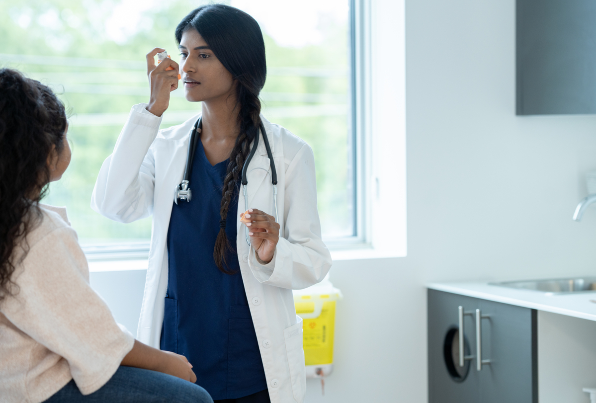 1356463622 A female doctor holds out an inhaler as she instructs her young patient on how to properly use it for her Asthma. The patient is dressed casually and sitting up on the table as she pays close attention to the instructions.